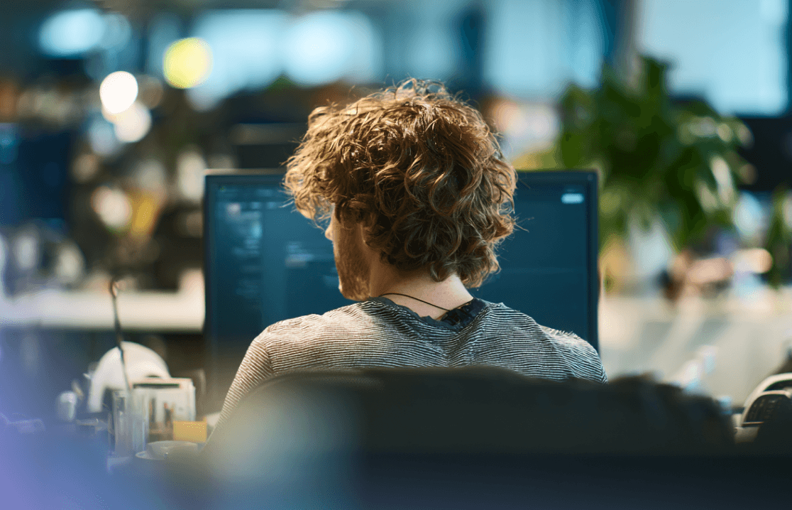 Blick über die Schulter auf eine:n Entwickler:in mit lockigem Haar, die oder der in einem modernen Büro an einem Computer arbeitet.